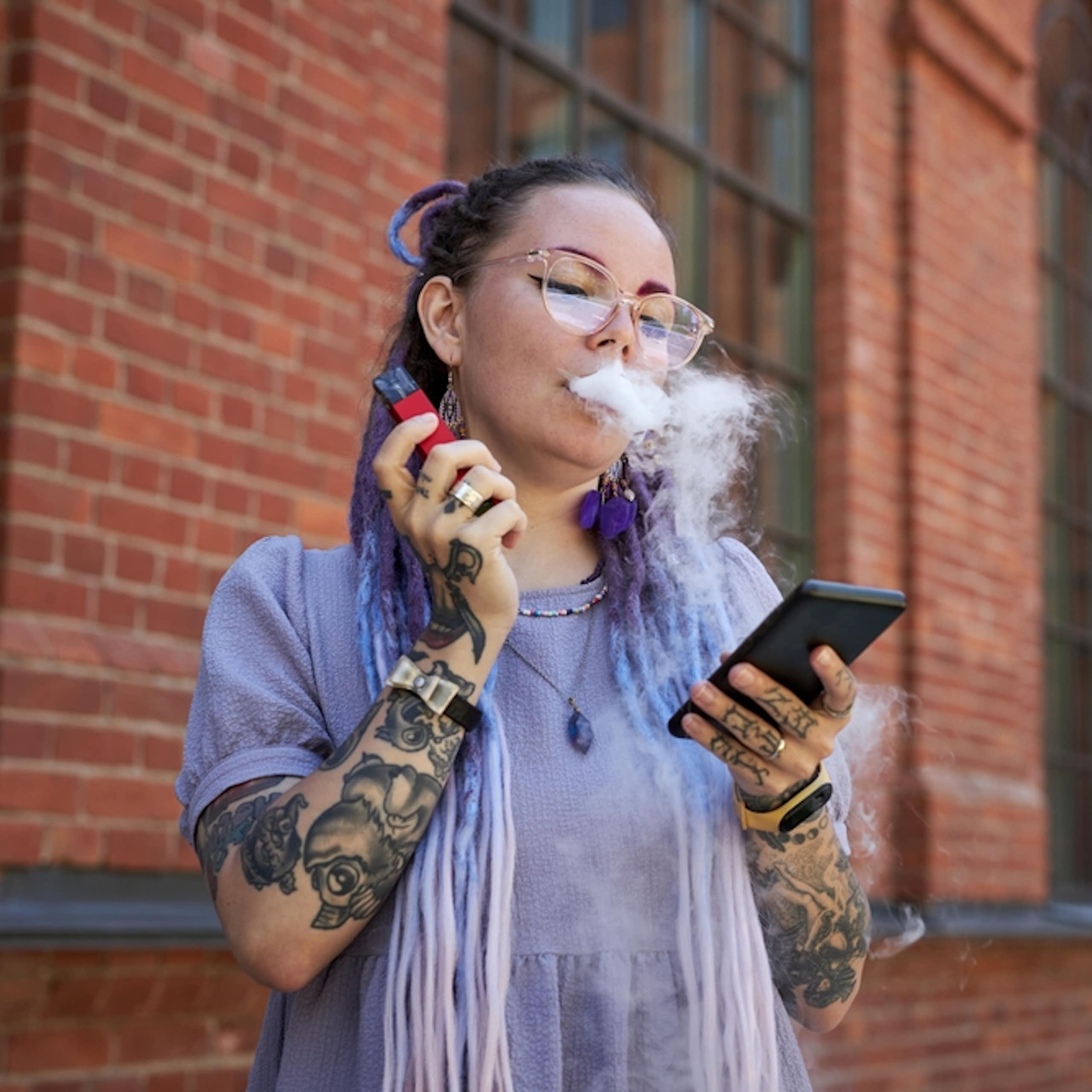 Young woman vaping, holding smartphone, outside redbrick building