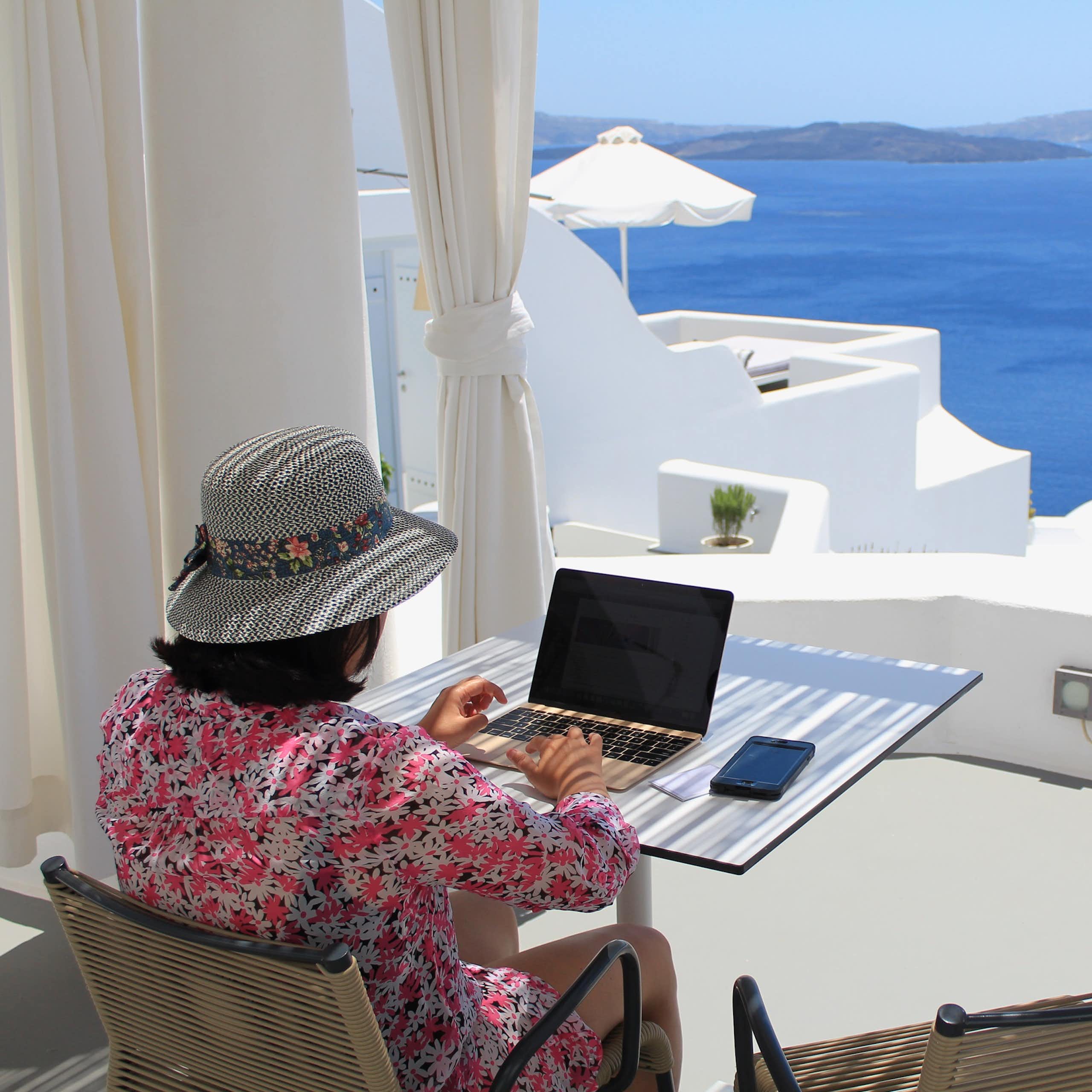 A woman works in her laptop by a window overlooking a Greek village.