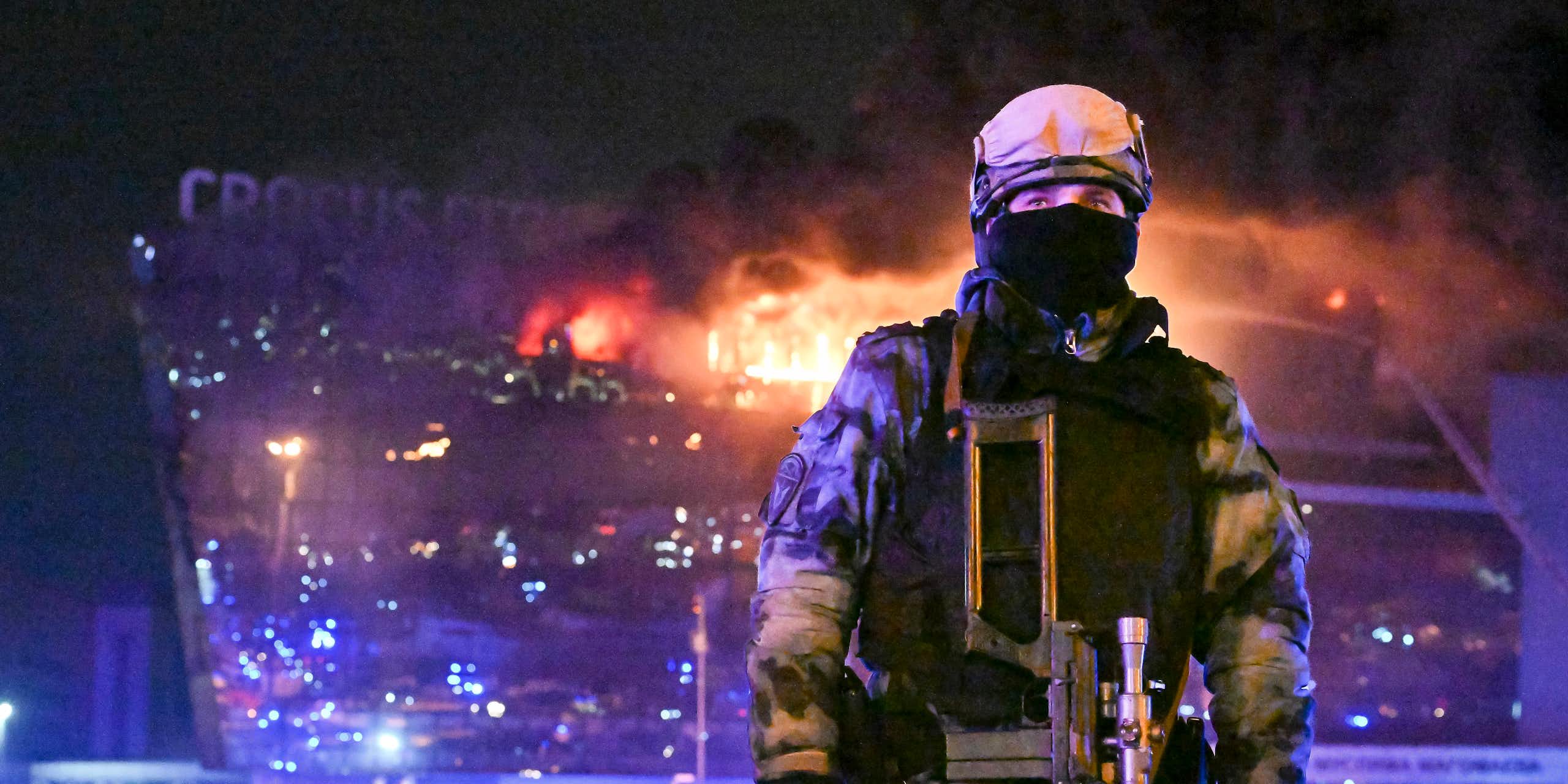 Un soldat se trouve au premier plan de la photo tandis qu'un bâtiment en flammes se trouve à l'arrière-plan.
